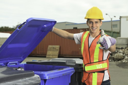 A young male worker wearing a yellow safety helmet, a neon orange high-visibility vest with reflective strips, and purple work gloves is standing outdoors near a row of large black wheelie bins, with his right hand on the open lid of a blue bin and giving a thumbs-up with his left hand. In the background, there are industrial buildings, a container, and a paved area, indicating a commercial waste collection environment. The scene is well-lit with natural daylight, and the worker has a friendly, professional expression. This image relates to rubbish removal services, illustrating a professional and efficient approach to waste management, potentially serving areas within the Forest Gate or wider London region, as suggested by the company name and location context.