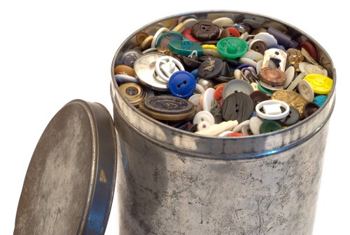 A round metal container, likely a rubbish bin, filled with a diverse assortment of plastic and metal buttons of various sizes, colours, and finishes. The buttons include both matte and shiny surfaces, with some appearing worn or aged, predominantly in shades of black, white, blue, brown, and green. The bin is situated outdoors on a paved or asphalt surface, with a slightly open, hinged lid placed on the ground beside it, revealing the densely packed contents inside. The environment suggests a setting related to waste collection or disposal, consistent with services provided by Commercial Waste Removal Forest Gate within the London postcode area. The background is not visible, focusing entirely on the container and its contents, emphasizing the variety of discarded buttons that might be part of household or manufacturing waste collected during rubbish removal operations in the Forest Gate area.