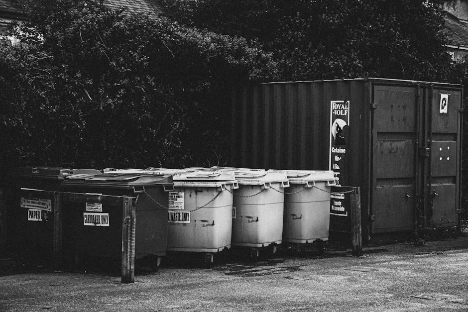 A collection of overflowing rubbish bins and scattered waste is situated on a paved urban pavement near a parking area. The scene includes a large grey mixed paper and card waste container with its li